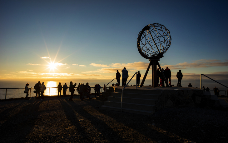 Globus-Skulptur am Nordkap in Norwegen bei Sonnenuntergang mit Touristen im Vordergrund.
