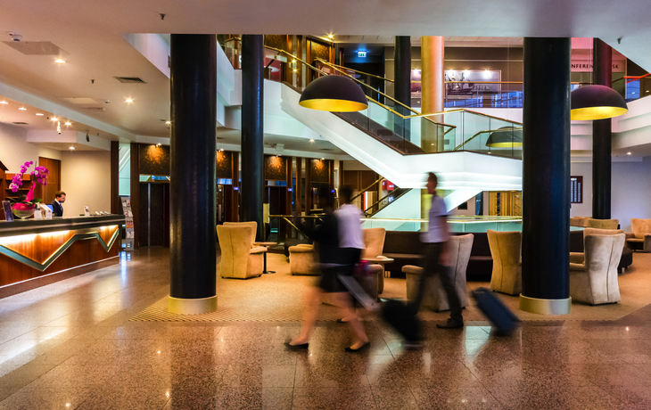 Hotel-Lobby mit Empfangstresen, zwei Personen mit Rollkoffern, moderne Einrichtung.