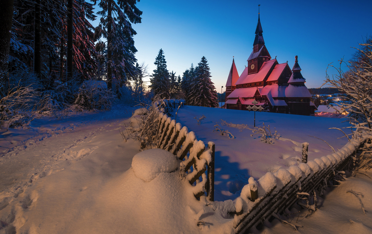 Stabkirche Hahnenklee im Schnee bei Abenddämmerung, umgeben von Bäumen.