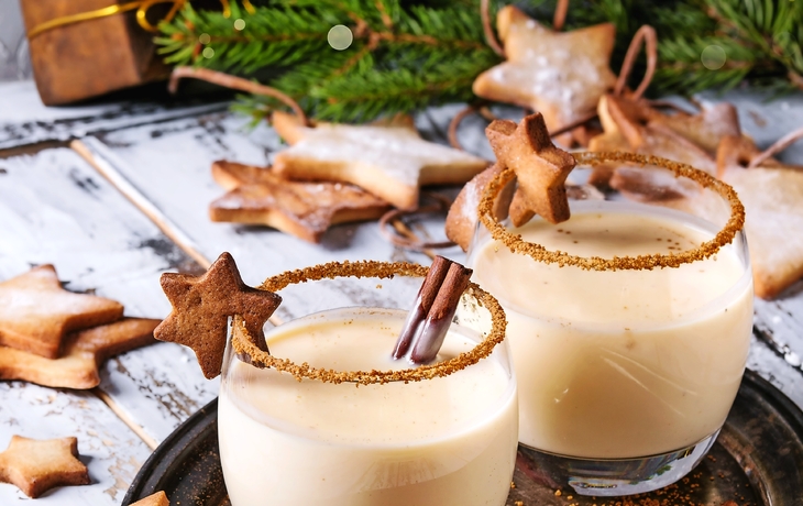 Zwei Gläser Eggnog mit Zimt und Sternkeksen auf einem Tablett, weihnachtlich dekoriert.