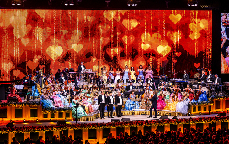 Orchester spielt vor Bühne mit Herzdekor, viele Künstler und bunte Kleider.