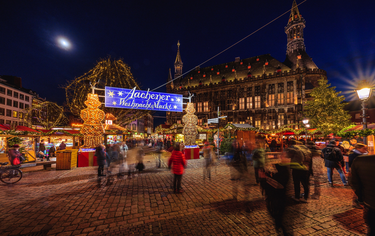 Eingang am Aachener Weihnachtsmarkt vor dem beleuchteten Rathaus in der Abenddämmerung.