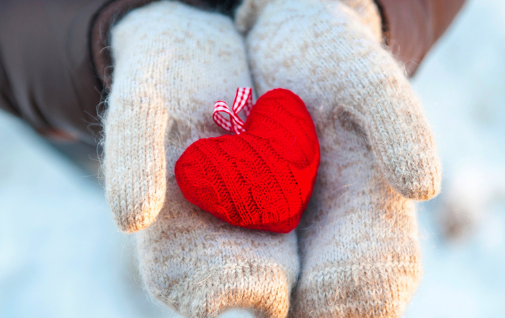 Hände in Handschuhen halten ein rotes Juleherz im Schnee.