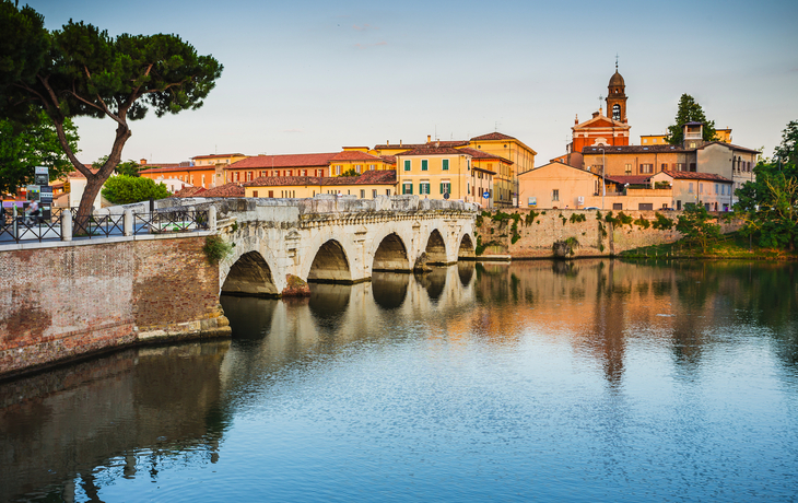 Historische Tiberiusbrücke in Rimini, Italien, mit reflektierendem Flussufer im Abendlicht.
