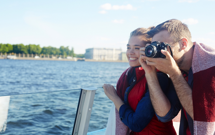 Paar auf Boot, Mann fotografiert, Wasser im Hintergrund.