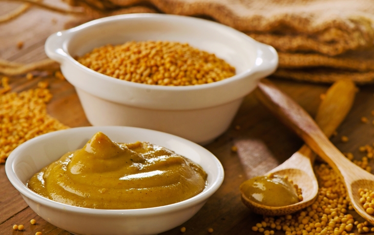 Senfkörner und Senfpaste in weißen Schalen auf einem Holztisch mit einem Holzlöffel.