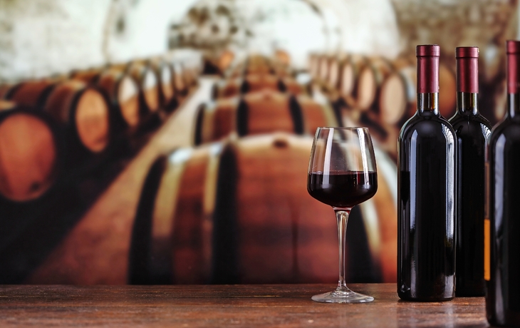 Weinflaschen und ein Weinglas in einem Weinkeller mit Fässern im Hintergrund.