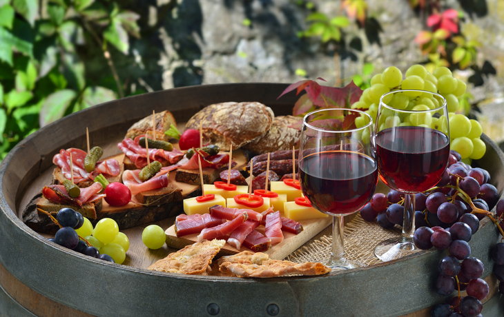 Wurstplatte mit Brot, Weintrauben und Rotweingläsern auf einem Fass.