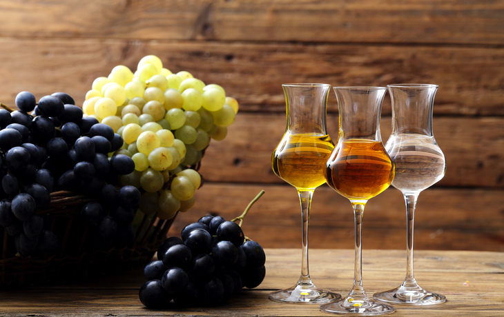 Drei Gläser Spirituosen und Trauben auf Holztisch vor Holzwand.