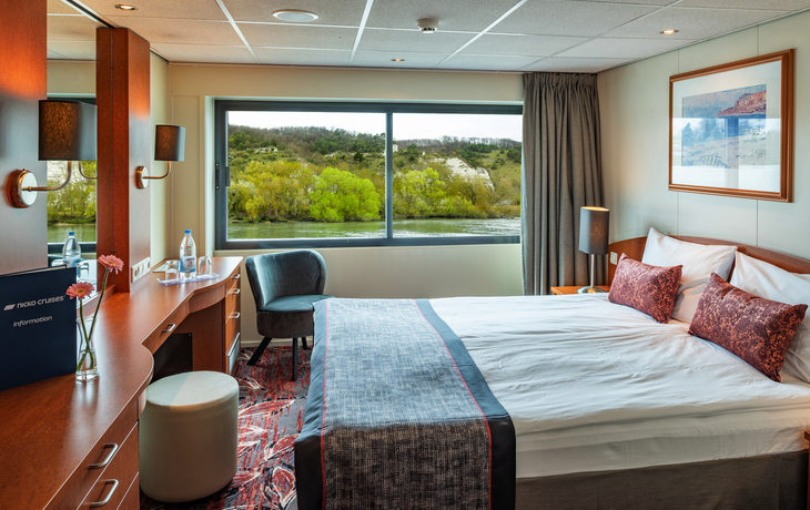 Kabine mit Doppelbett, Schreibtisch und großem Fenster mit Aussicht auf Flusslandschaft.