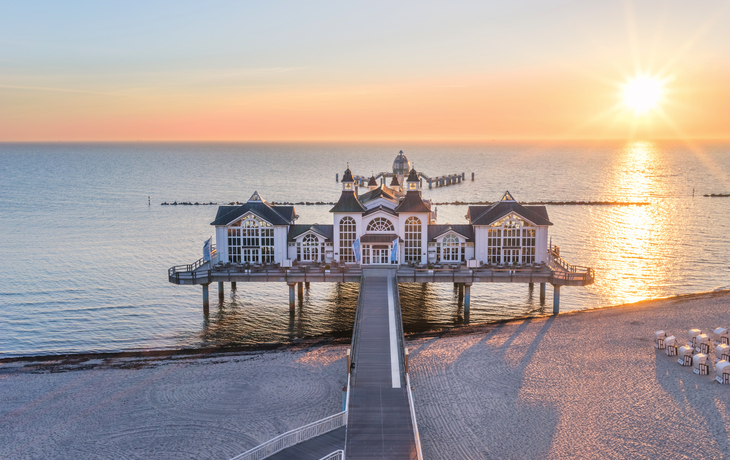 Sonnenaufgang über der Seebrücke in Sellin, Ostsee, mit Sandstrand im Vordergrund.