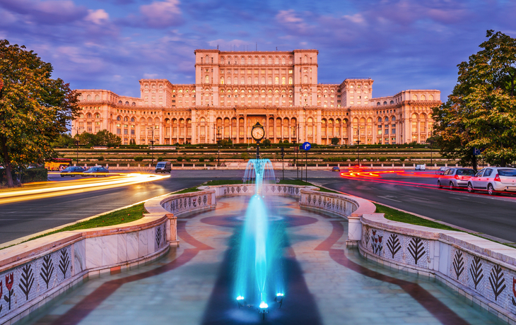 Parlamentspalast Bukarest bei Abenddämmerung, mit Brunnen und vorbeifahrenden Lichtern.