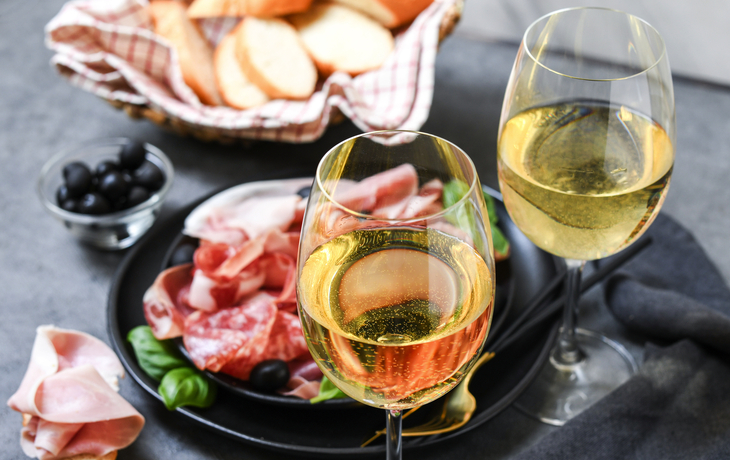 Zwei Gläser Weißwein, Brot, Schinken und Oliven auf einem Tisch.