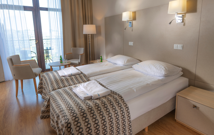 Helles Hotelzimmer mit zwei Betten, Tisch, Stühlen und Balkonzugang.