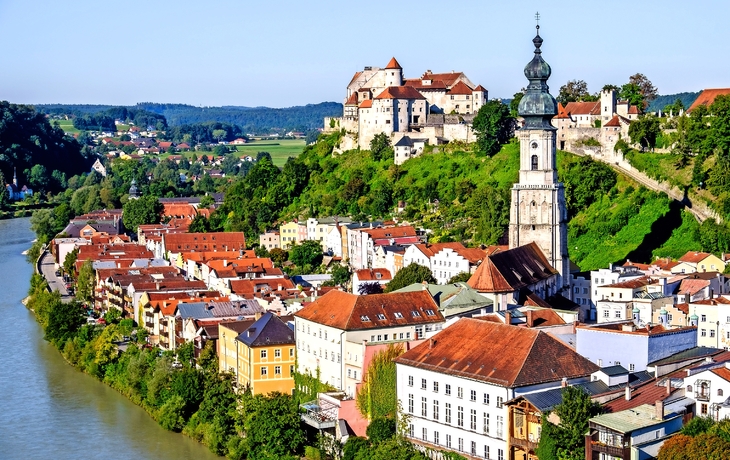 Stadtansicht mit historischen Gebäuden, Fluss und Burg auf einem Hügel.