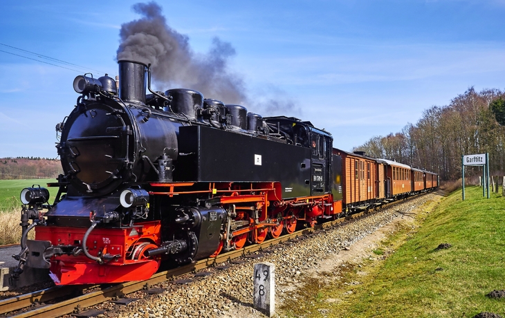 Dampflokomotive zieht einen historischen Zug durch eine ländliche Landschaft.