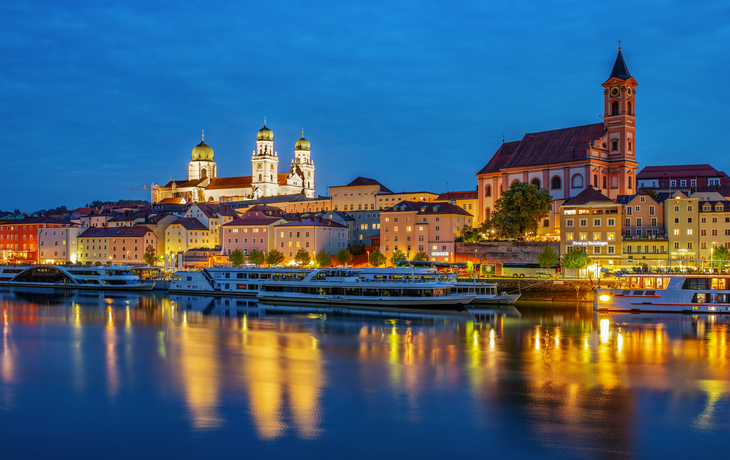 Abendliche Stadtansicht mit beleuchteten Gebäuden und Fluss im Vordergrund.