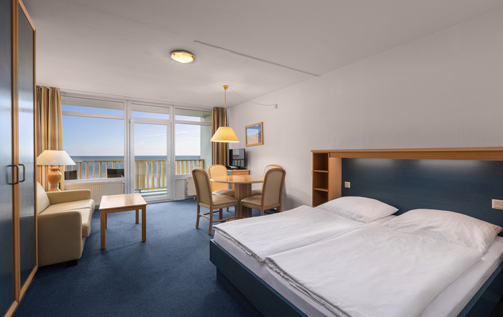 Ferienwohnung mit Doppelbett, Sofa, Tisch, Stühlen und Meerblick durch große Fenster.