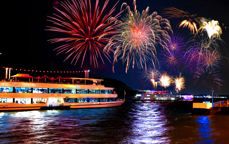 Feuerwerk über dem Rhein mit beleuchteten Kreuzfahrtschiffen bei Nacht.