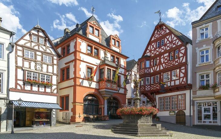 Fachwerkhäuser an einem Platz mit Brunnen und blauem Himmel.
