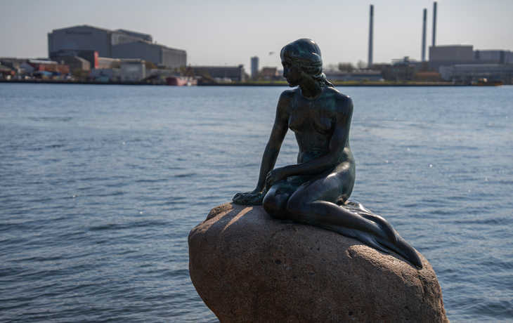 Bronzestatue einer Meerjungfrau auf einem Felsen im Wasser mit Industriekulisse.
