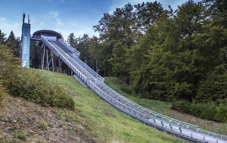 Skisprungschanze in bewaldeter Umgebung bei klarem Himmel.